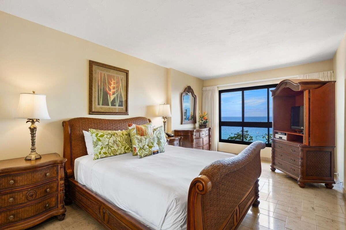 A spacious bedroom with a large bed, wooden furniture, a television, and a view of the sea.