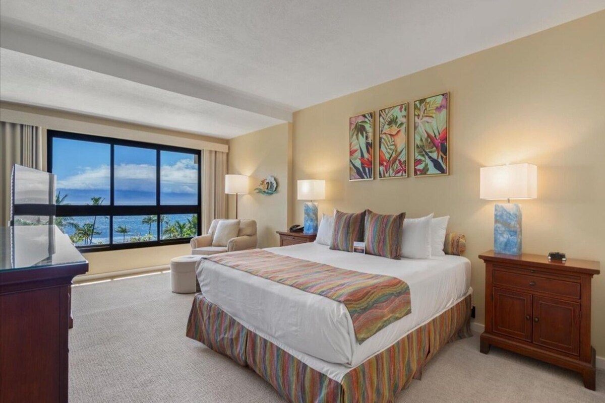 A hotel room with a large bed, two bedside tables, a desk, a chair, a window with a view of the ocean, and three wall-mounted paintings.