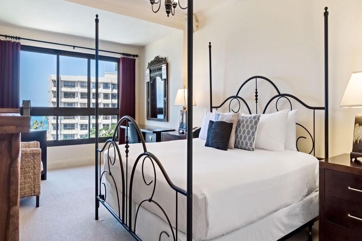 A four-poster bed with a white mattress and decorative pillows, a wooden nightstand, a desk with a chair, and a large window with curtains.
