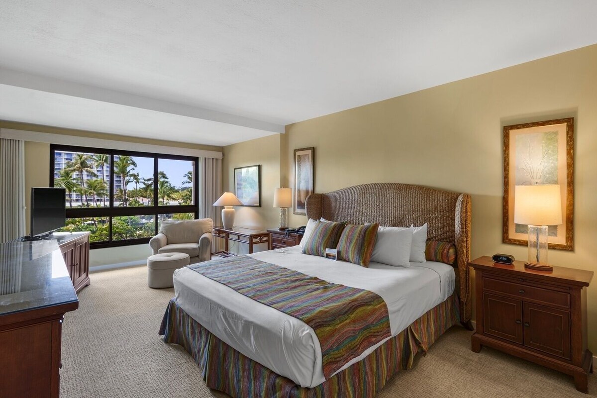 A hotel room with a large bed, a desk, a chair, a TV, and a window with a view of palm trees.