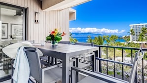 A balcony with a table and chairs, a vase with flowers, and a view of the ocean and palm trees. - Kaanapali Alii-2 Bedroom Partial Ocean Views (Lahaina)
