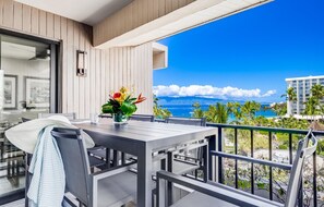 A balcony with a table and chairs, a vase with flowers, and a view of the ocean and palm trees. - Kaanapali Alii-2 Bedroom Partial Ocean Views (Lahaina)