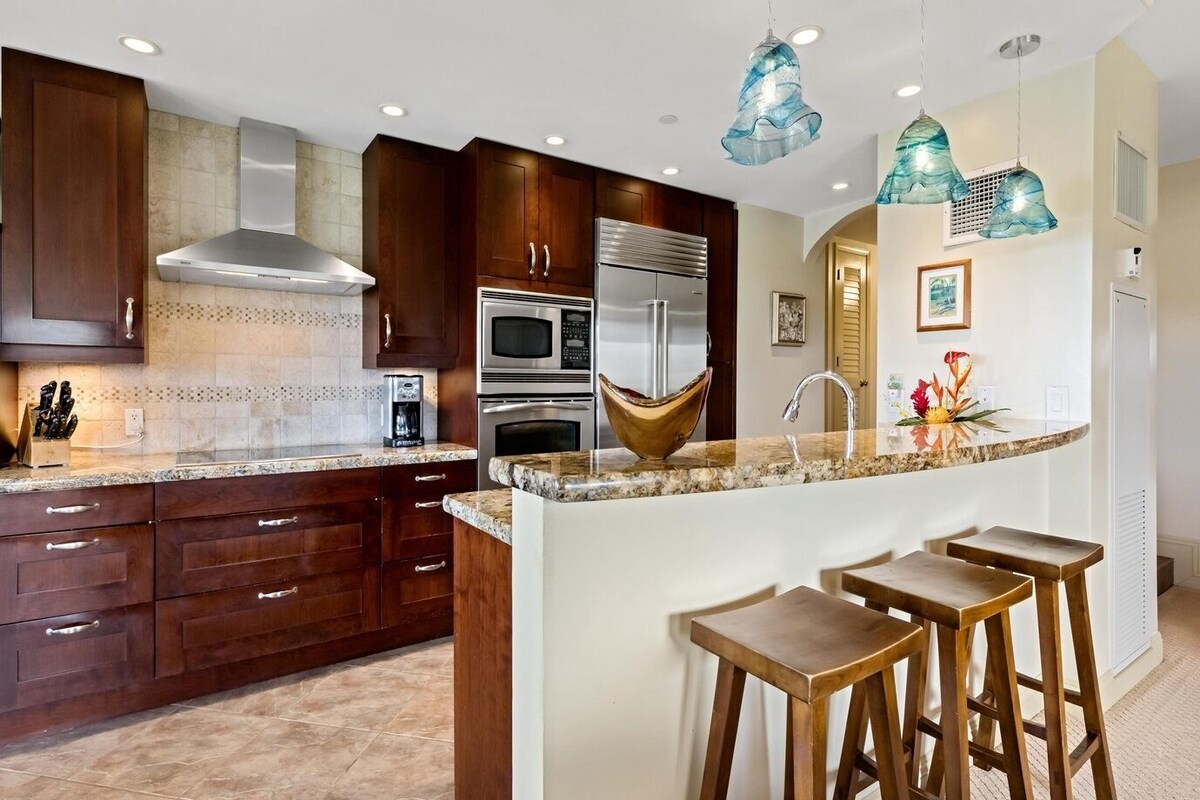 A modern kitchen with dark wooden cabinets, stainless steel appliances, a marble countertop, and a central island with stools.