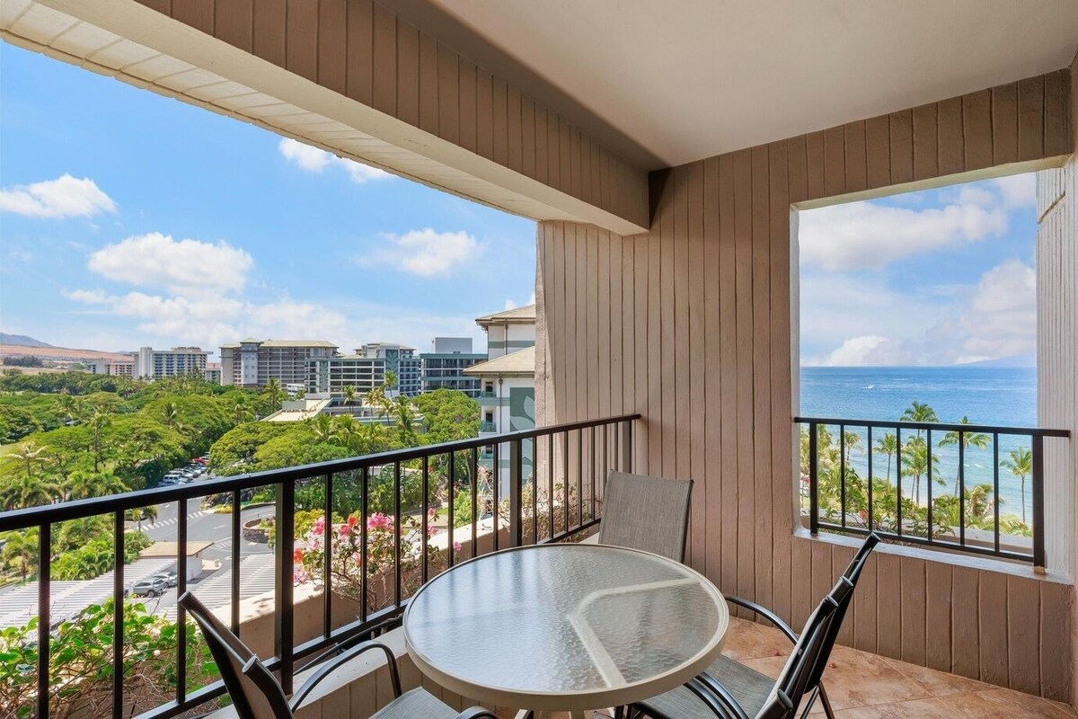 A balcony with a round table and chairs, a view of the ocean, and buildings in the background.