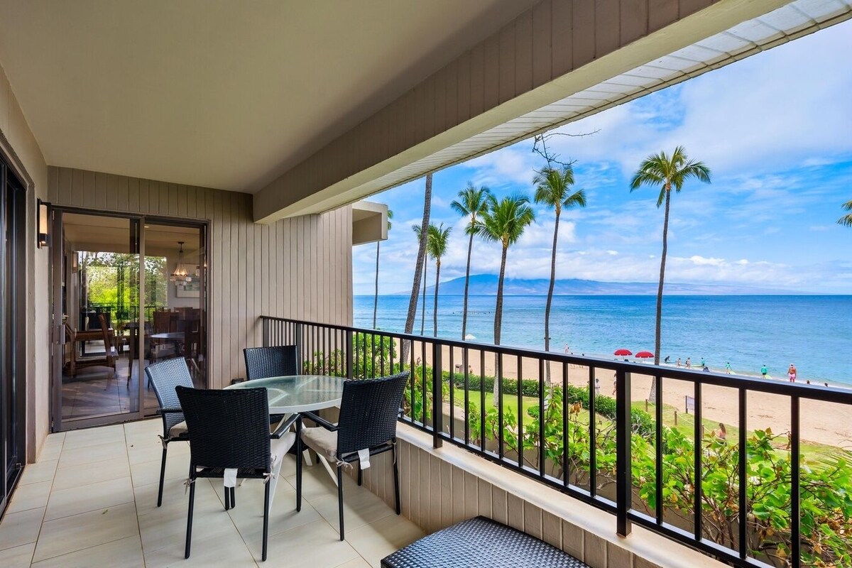 Ein Balkon mit Glastisch und Stühlen, der Blick auf einen Strand mit Palmen und den Ozean gewährt.