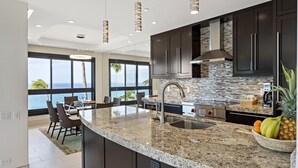 A modern kitchen with dark cabinets, a granite countertop, and a view of the ocean. - Kaanapali Alii-2 Bedroom Ocean Front Views (Lahaina)