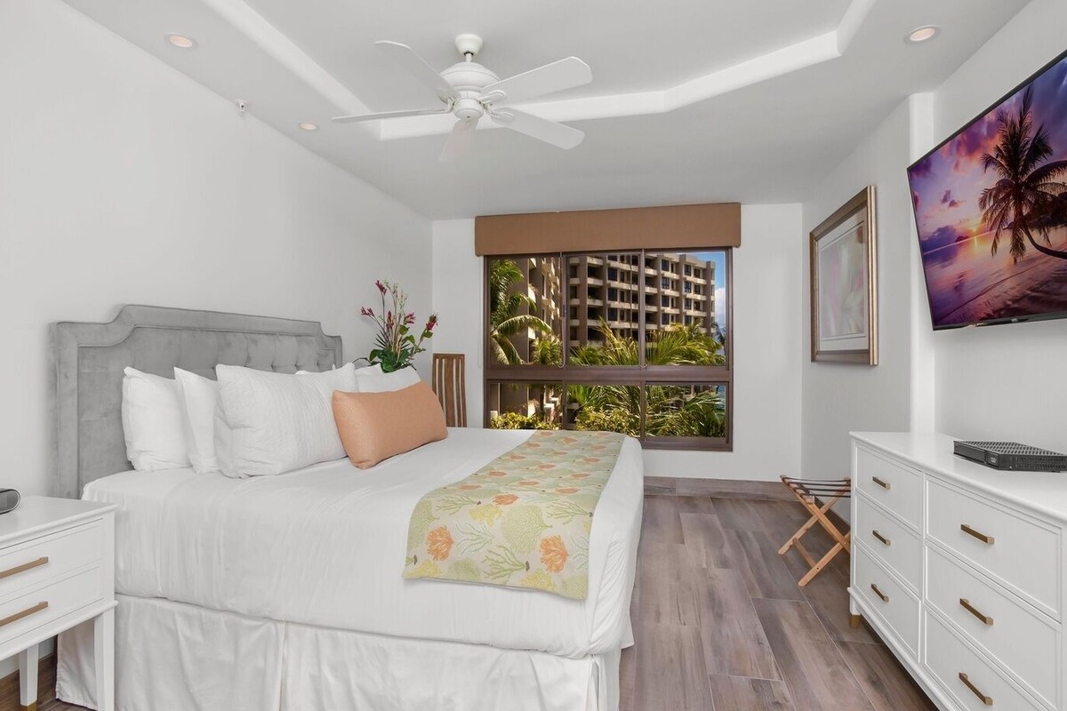 A bedroom with a bed, bedside tables, a TV, a ceiling fan, a window with a view of a building, and a plant.