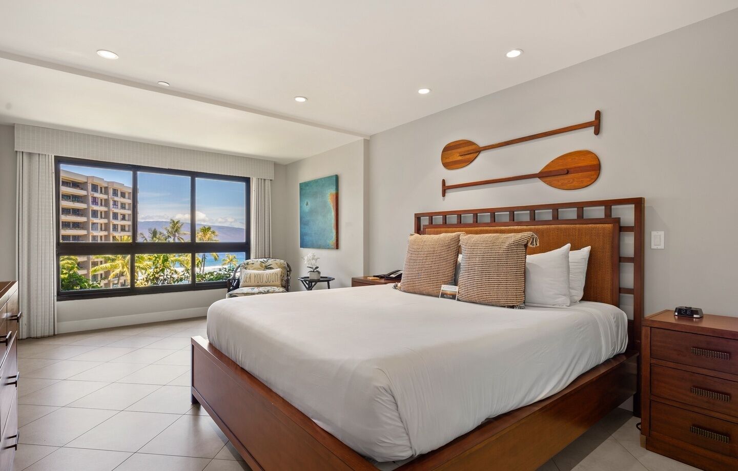 Une chambre d’hôtel avec un grand lit, une vue sur des palmiers et un immeuble par la fenêtre, une tête de lit en bois et une table de chevet avec une horloge.