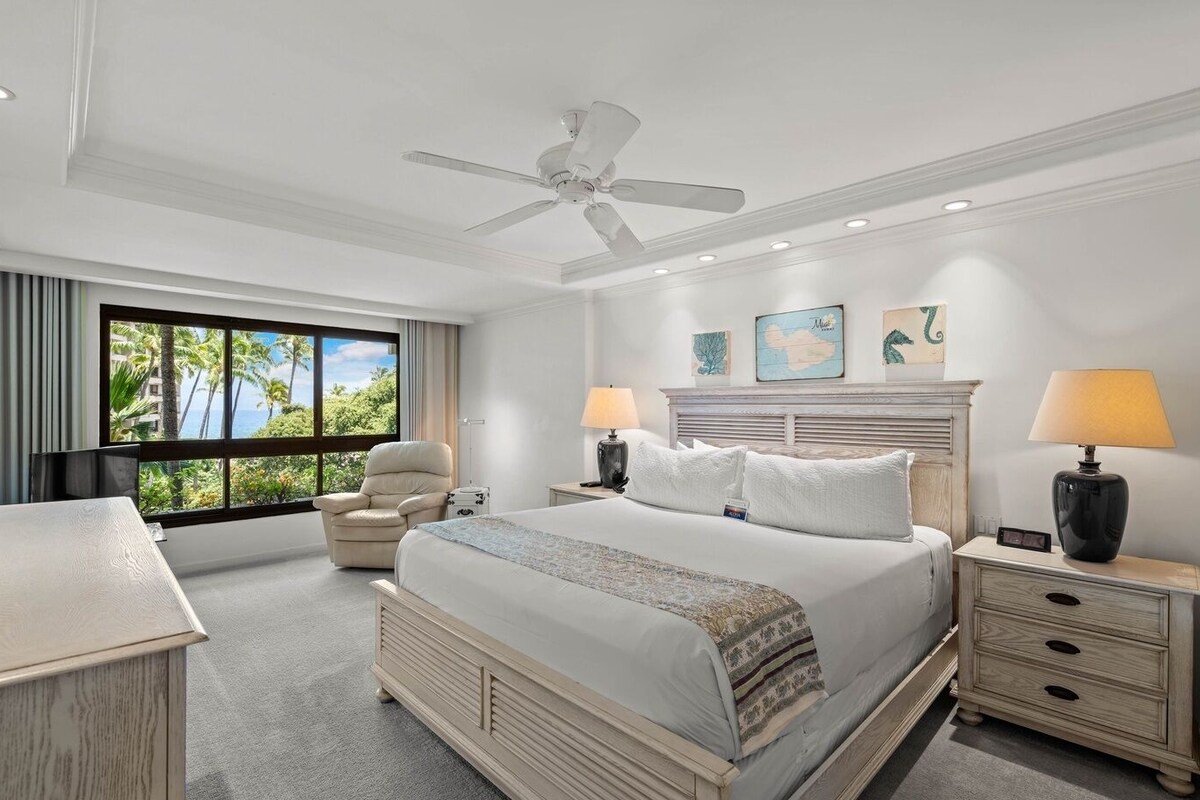 A hotel room with a bed, two bedside tables, a chair, a desk, a ceiling fan, and a large window with a view of palm trees.