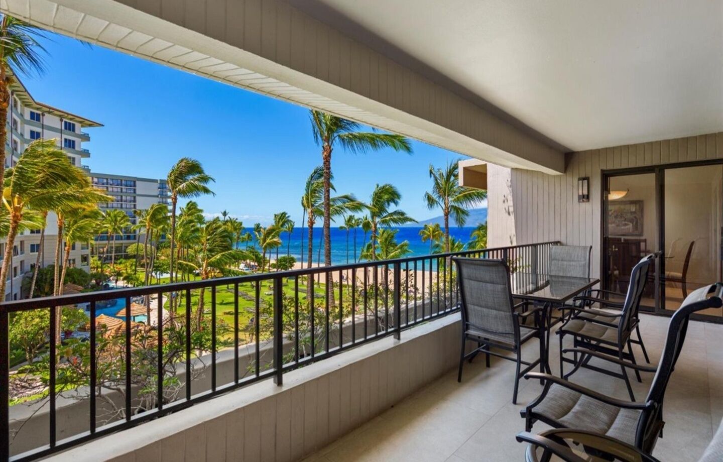 A balcony with a view of the ocean and palm trees, a dining table with chairs, and a pool in the background.