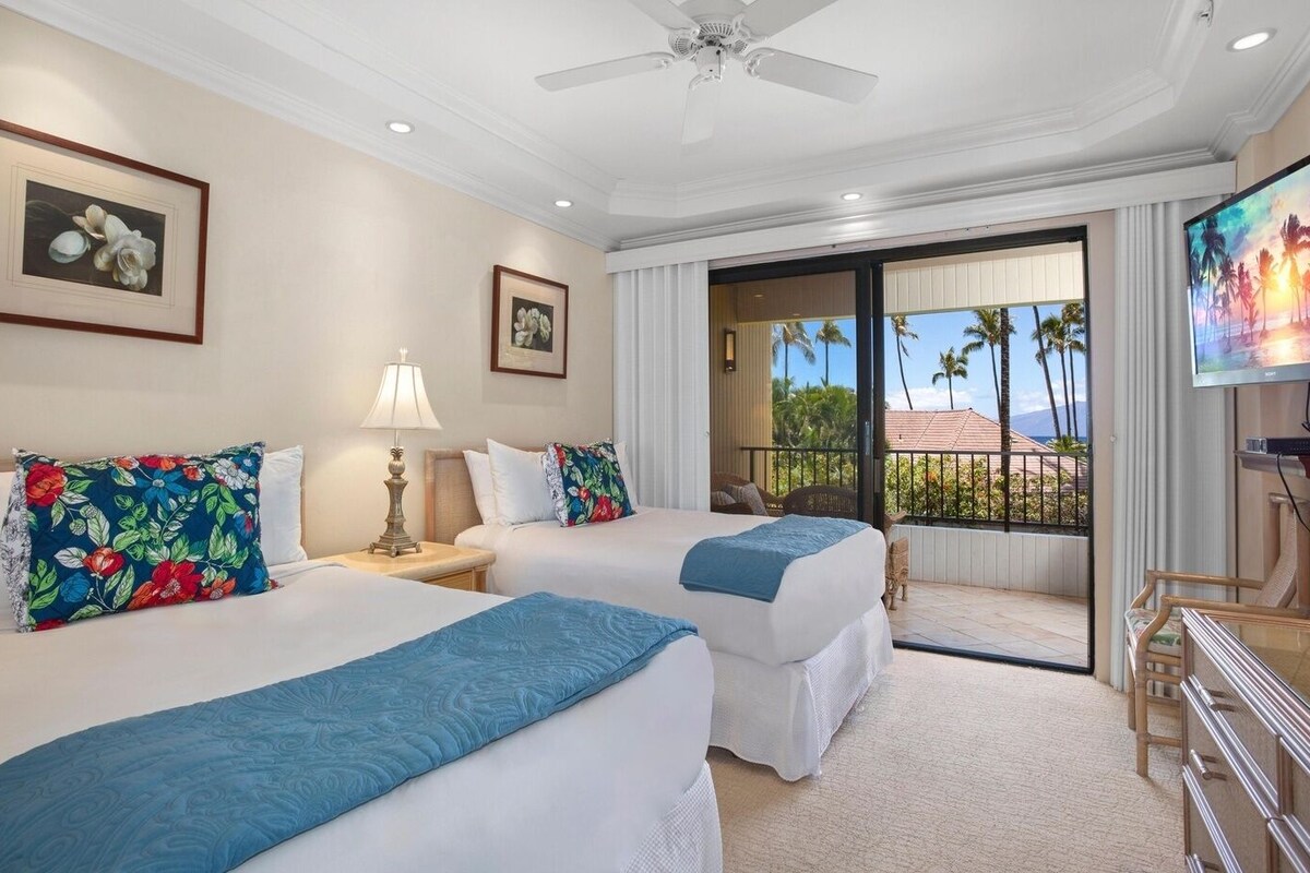 A hotel room with two beds, a ceiling fan, a TV, and a balcony with a view of palm trees.