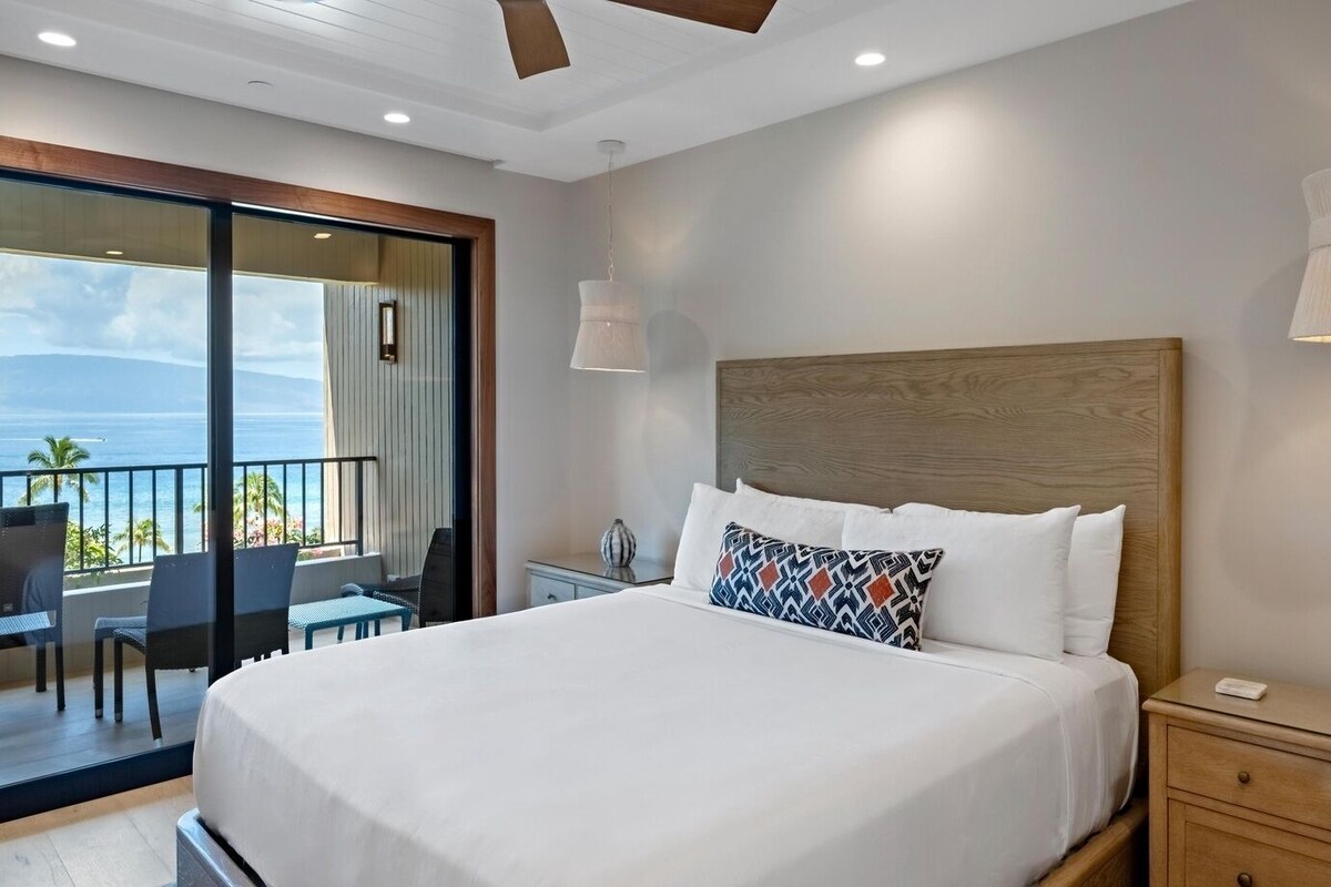 A hotel room with a large bed, a nightstand, a desk with chairs, and a balcony with a view of the ocean.