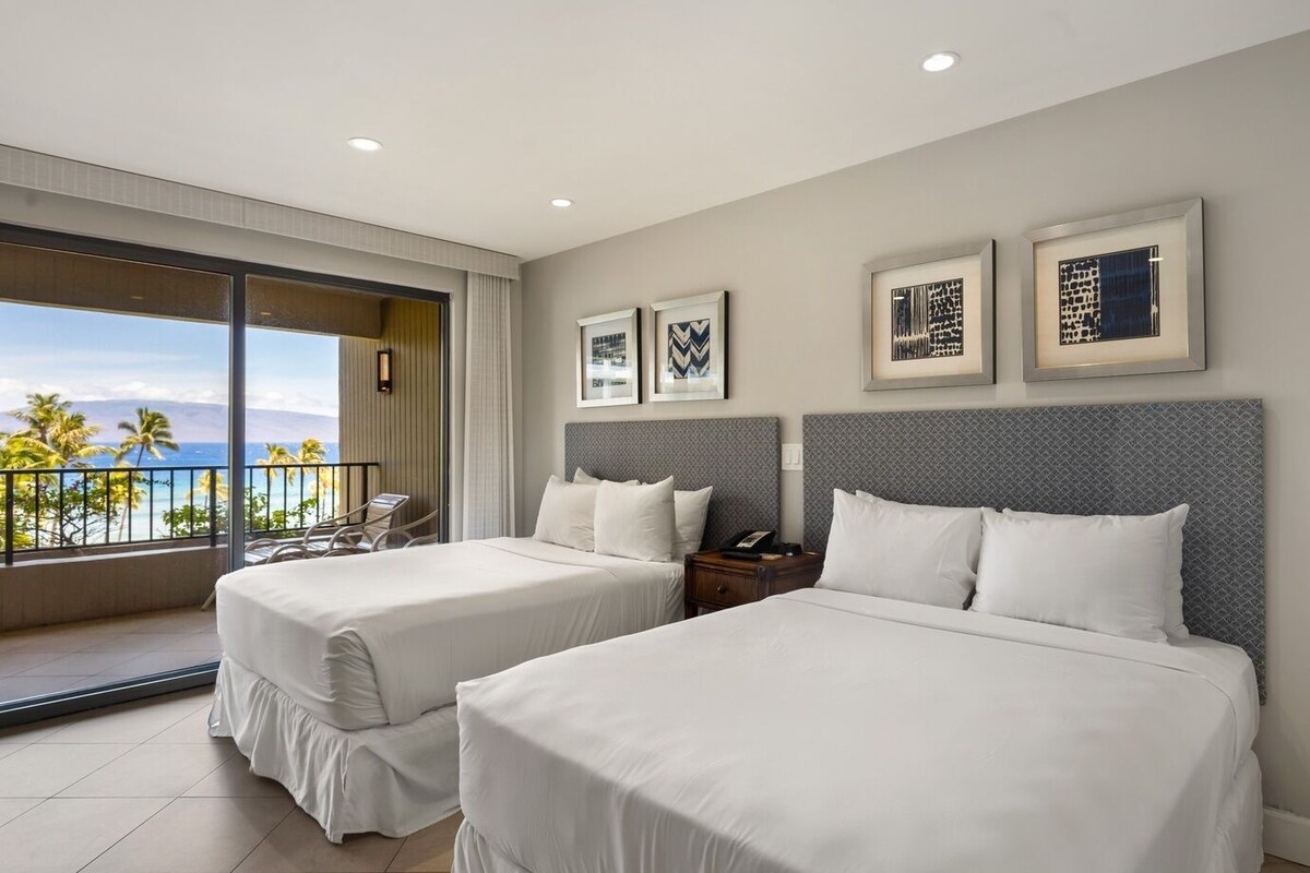 A hotel room with two beds, a balcony with a view of the beach, and framed artwork on the wall.
