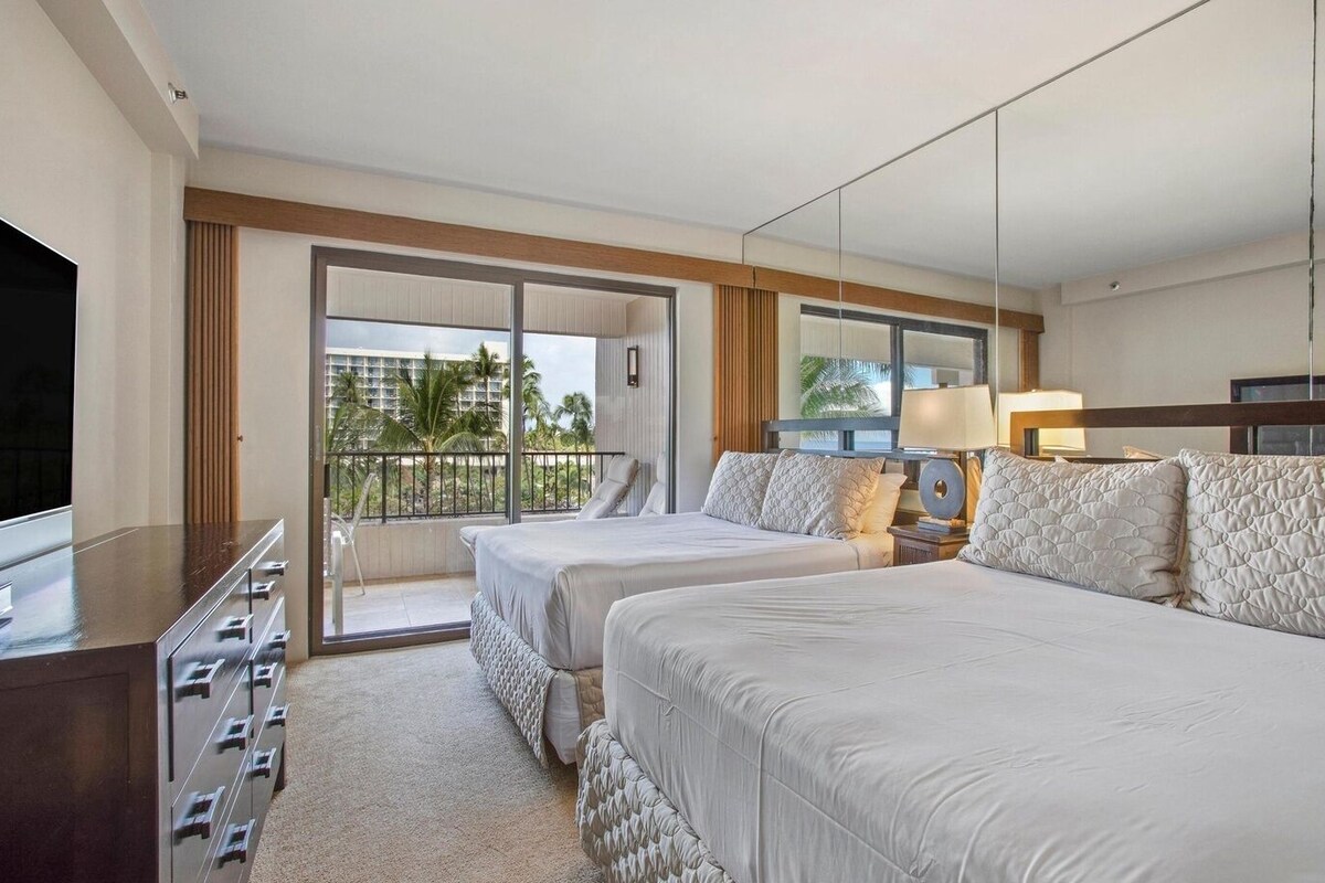 A hotel room with two beds, a TV, a desk with drawers, a balcony with a view, and a window with blinds.