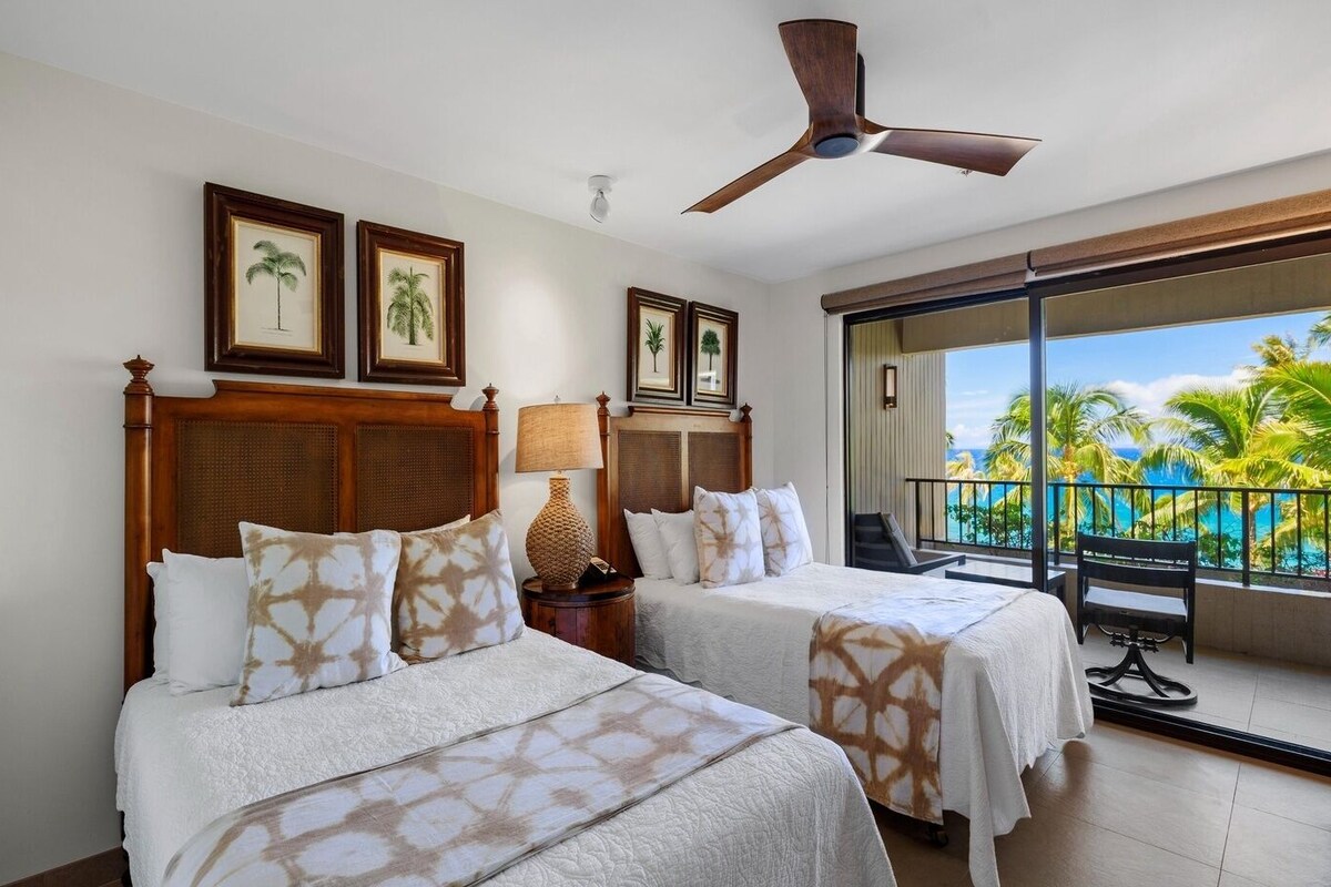 A hotel room with two beds, a ceiling fan, a large window with a view of palm trees and the ocean, and framed pictures on the wall.