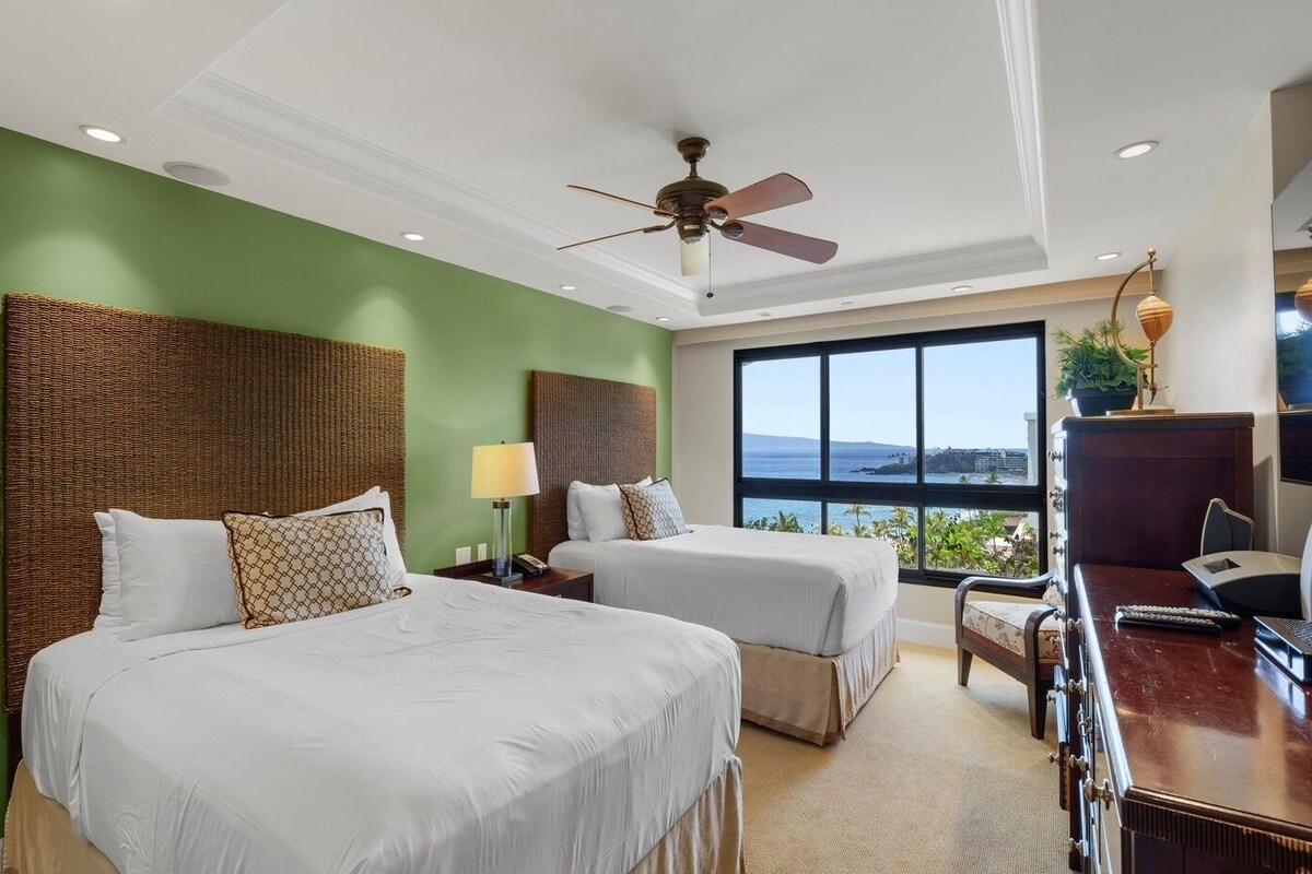 A hotel room with two beds, a desk with a chair, a desk lamp, a ceiling fan, and a large window offering a view of the ocean.
