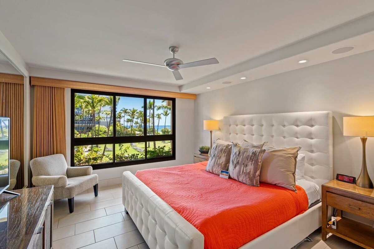 A hotel room with a large bed, a chair, a desk, a ceiling fan, and a view of palm trees.