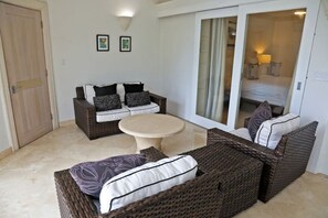 Una habitación con un conjunto de sofá de mimbre, una mesa redonda y un ventanal corredero que da a un dormitorio.