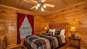 Ein Schlafzimmer einer Holzhütte mit einem Bett, zwei Nachttischlampen, einem Deckenventilator und einem Fenster mit roten Vorhängen.