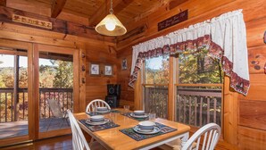 A wooden cabin interior with a dining table set for four, a large window with curtains, and a hanging light fixture. - Hawks Nest #1907 (Sevierville)