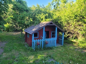Eine kleine, rote Holzhütte mit einer blauen Tür und Fenstern, umgeben von Grünpflanzen.