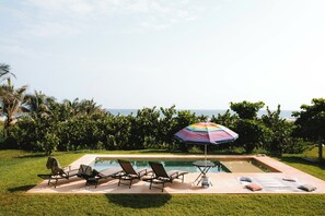 A swimming pool with lounge chairs and an umbrella, surrounded by greenery and a view of the ocean. - Beach house with pool El Cortijo (Puerto Escondido)