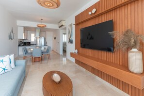 A modern living room with a flat-screen TV mounted on a wooden wall, a dining area with a glass table and chairs, and a kitchen in the background. - Exquisite apt : Private beach : Swimming pool : 2BR (Punta Cana)