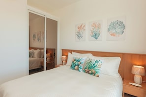 A neatly made bed with a white comforter and two decorative pillows, a wooden headboard, a nightstand with a lamp, and wall art depicting sea plants. - Exquisite apt : Private beach : Swimming pool : 2BR (Punta Cana)