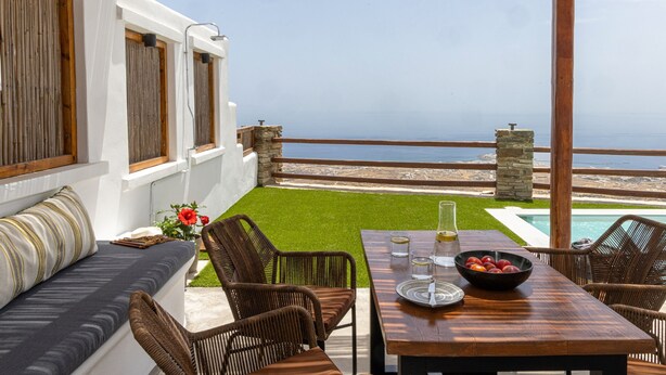Une terrasse avec une table en bois, des chaises en osier, un banc rembourré et une vue sur la mer.