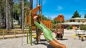 Leikkikenttä, jossa on liukumäki ja kiipeilytelineitä, ja jonka ympärillä on puita ja rakennus.