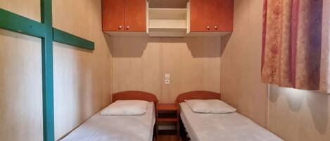 A small, compact room with two single beds, wooden cabinets above, and a curtain on the right.