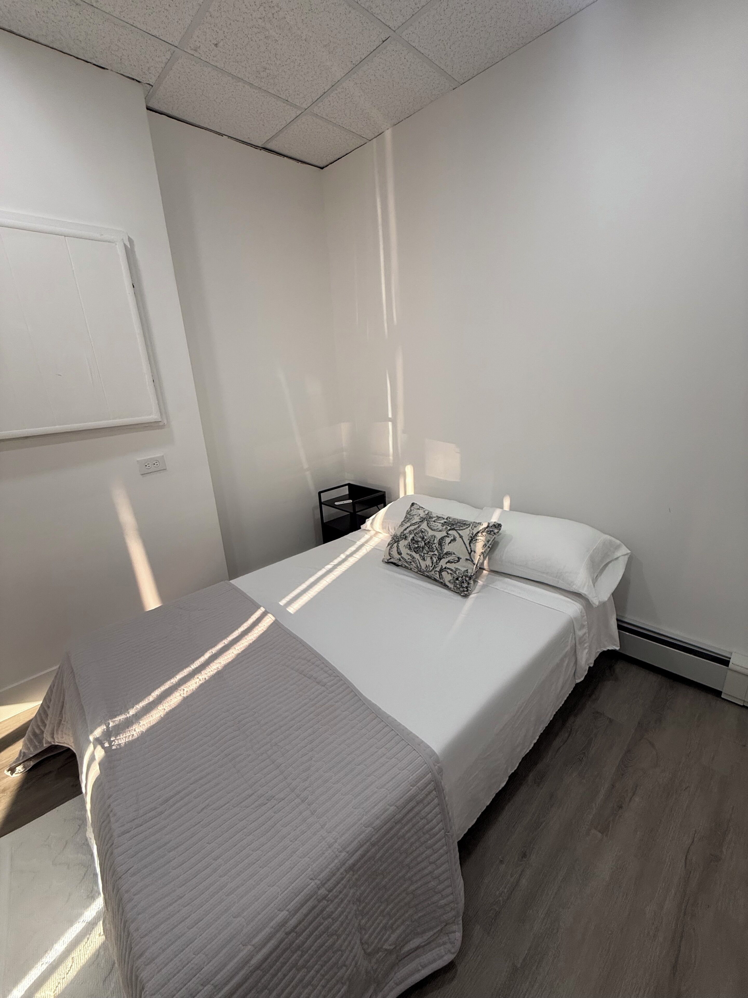A hotel room with a bed, a bedside table, a nightstand, and a wall-mounted light fixture.