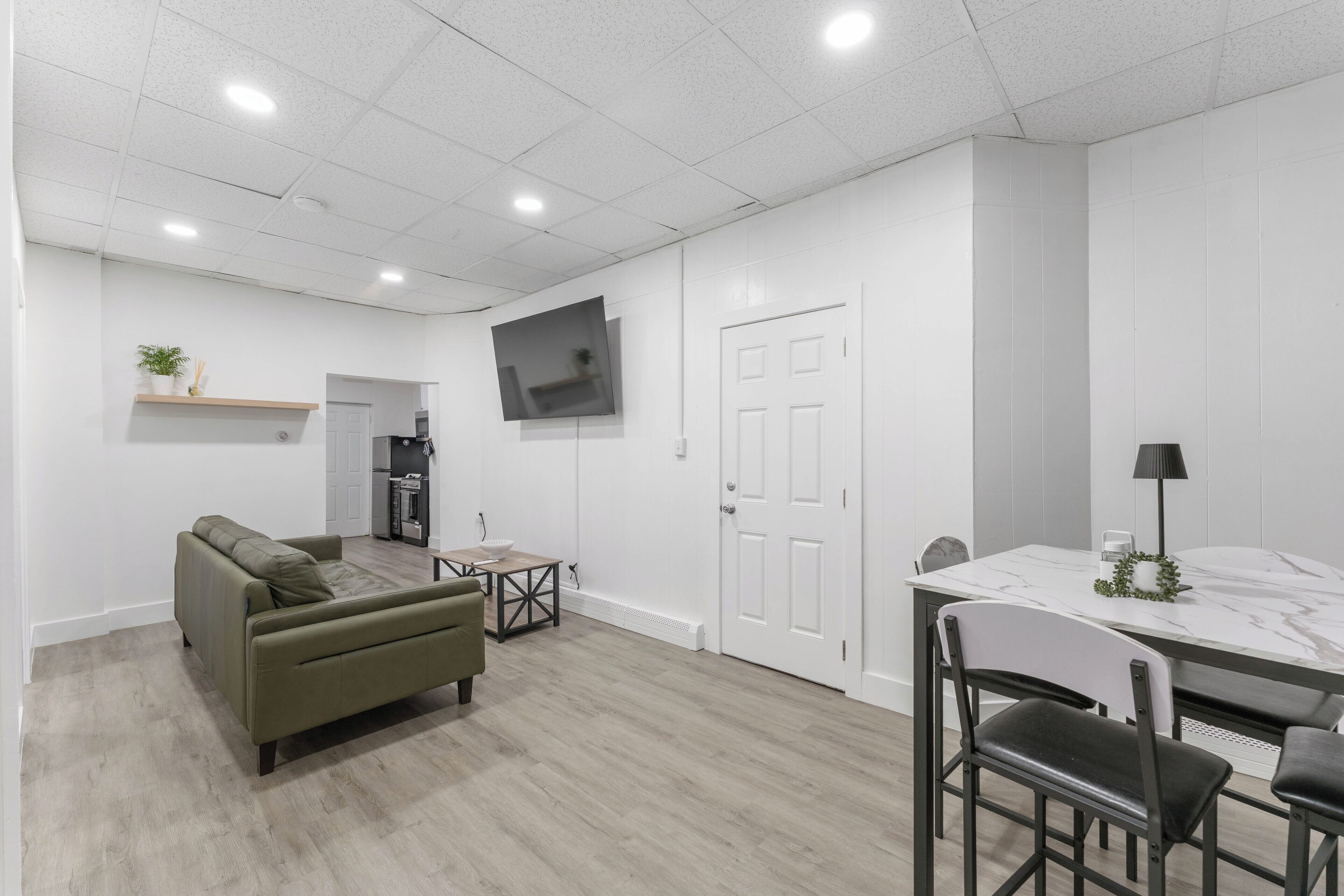 A modern living room with a sofa, a dining table with chairs, a TV mounted on the wall, and a door.