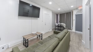 A modern living room with a large flat-screen TV, a wooden coffee table, a green sofa, and a dining area with a table and chairs. - Spacious 3-bedroom apartment in gorgeous West New York with AC (West New York)