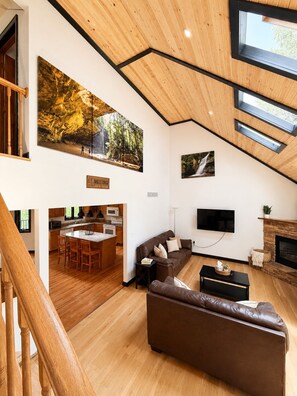 A spacious living area with a wooden ceiling, a large painting of a forest, a fireplace, and a modern kitchen visible through an open door. - Jake’s Lodge w/ Hot Tub,  Sleeps 14 , located in Hocking Hills (Logan)