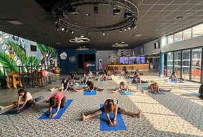 Ein großzügiger Innenbereich mit einer Gruppe von Menschen, die Yoga auf Matten praktizieren, einer Bühne mit Beleuchtungstechnik und einer Wand mit einem floralen Wandgemälde.