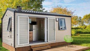 Een stacaravan met een openstaande deur, die een keuken, een klein terras en een groene stoel laat zien.