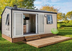 Ein Mobilheim mit geöffneter Tür, die eine Küche, eine kleine Terrasse und einen grünen Stuhl zeigt.