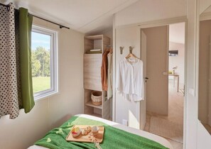 A bedroom with a bed, a window with curtains, a wooden shelf with clothes, and a tray with food. - Camping 4 étoiles - Parc aquatique - ccbeifa (Saint-Hilaire-de-Riez)