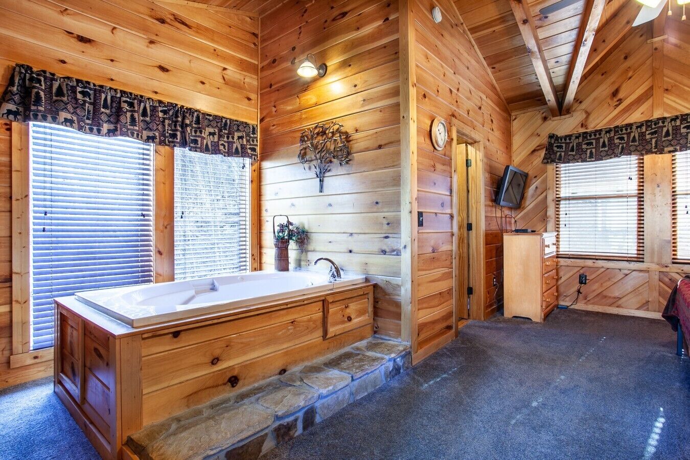 Interno di una baita di legno con una grande vasca da bagno in legno, un camino in pietra e un televisore montato a parete.
