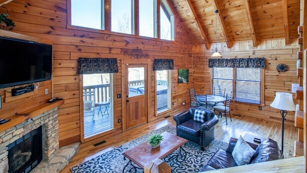 A log cabin interior with a stone fireplace, a television, a dining area, and a balcony.