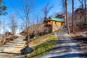 A wooden cabin with a green roof, a stone foundation, and a balcony overlooking a forested area.