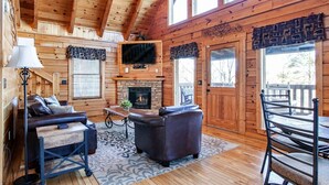 A log cabin interior with a fireplace, a television, a sofa, a dining table, and a view through the door.