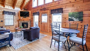 A cozy cabin interior with a fireplace, wooden walls, a dining area, and a large window.