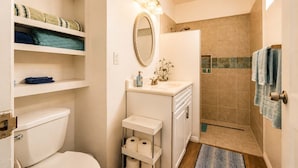 Une salle de bain équipée de toilettes, d’un lavabo et d’une douche. On y trouve des étagères avec des serviettes et un miroir au-dessus du lavabo.
