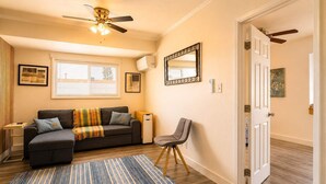 Un salon comprenant un canapé, un fauteuil, un ventilateur de plafond, une fenêtre avec des stores et une porte donnant sur une autre pièce.