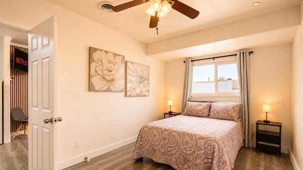 Une chambre à coucher comprenant un lit, deux tables de chevet, un ventilateur de plafond, une fenêtre avec des rideaux et des œuvres d’art murales.