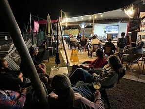 Ein Sitzbereich im Freien mit Tischen und Stühlen, eine erhöhte Plattform mit einer Leinwand und ein eingezäunter Bereich mit einem gelben Warnschild.