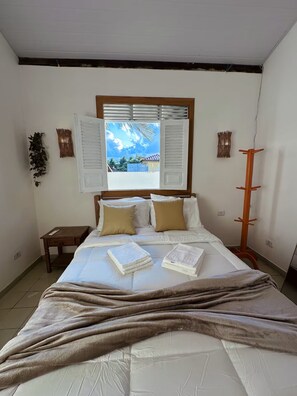 Ein Schlafzimmer mit einem Bett, einem Nachttisch, einem Fenster mit Fensterläden und Blick auf ein blaues Gebäude.