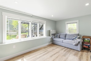 Ein Zimmer mit grauem Sofa, Holzboden und großen Fenstern mit Blick auf einen Hinterhof.